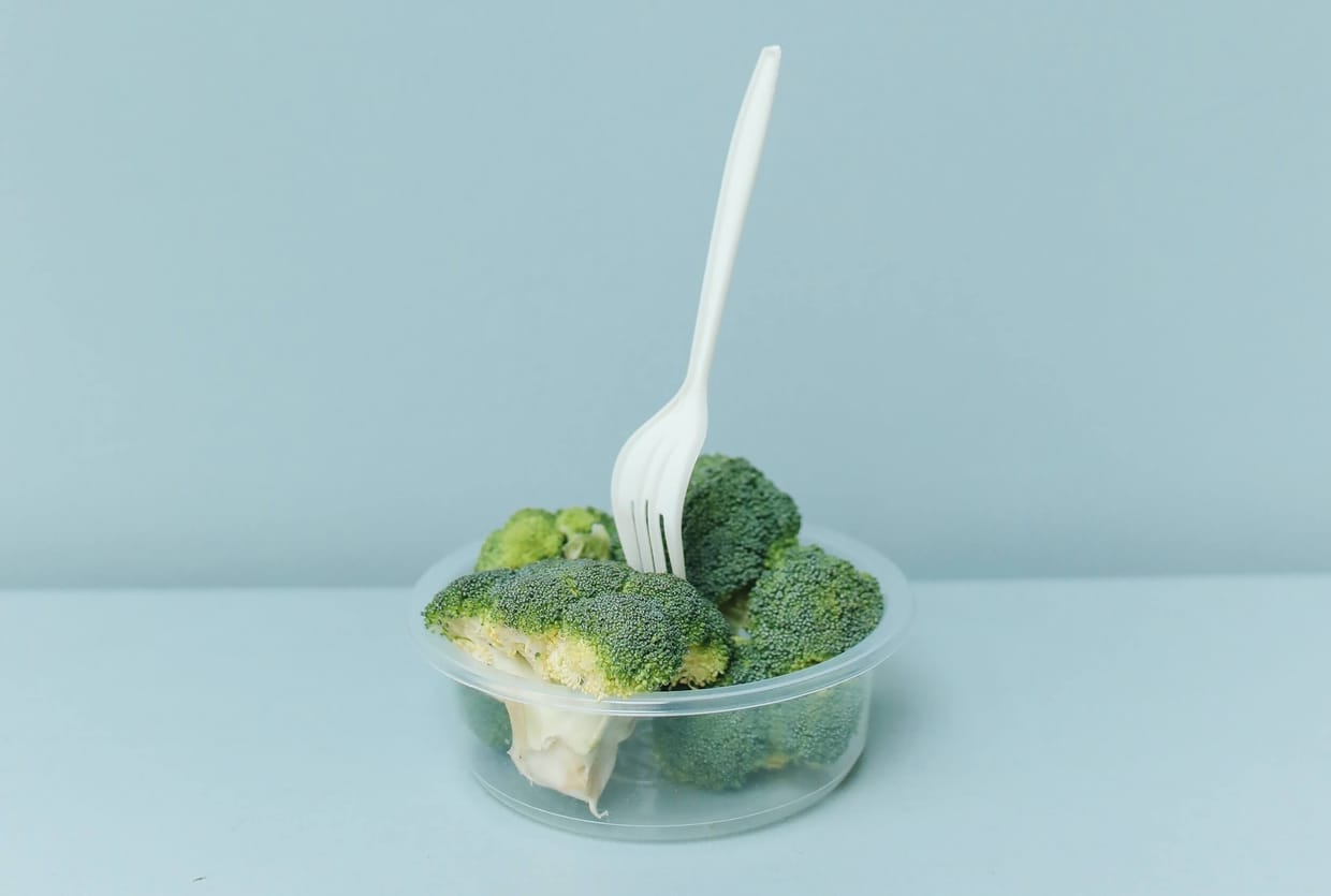 A clear plastic container filled with broccoli with a white plastic fork sticking out of it.