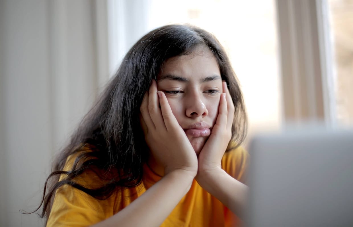 Young woman with dark hair looking bored while resting her head in her hands and looking at a laptop.